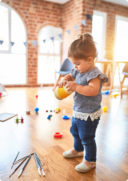 A young child stands on a wooden floor holding a yellow cup, surrounded by scattered toys and paintbrushes in a bright, sunlit room with brick walls and large windows.
