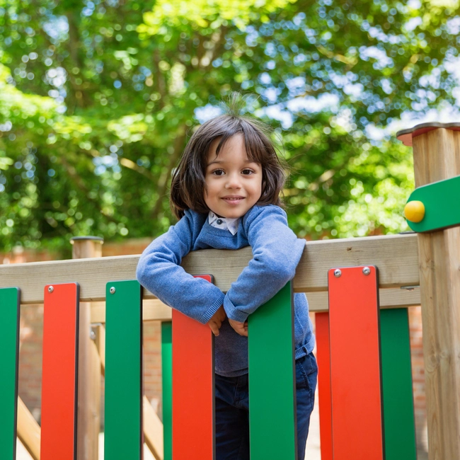 Little girl standing on the bridge of a colorful play set, smiling at the camera.