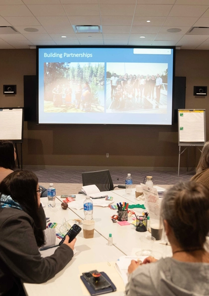A group of people sit at round tables in a conference room, listening to a presenter standing by a screen displaying a slide titled "Building Partnerships." Supplies and papers are scattered on the tables.