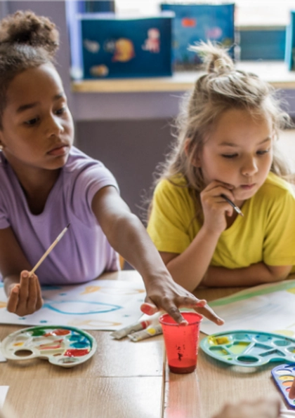 Two young children sit at a classroom table painting with watercolors. One child reaches for a paint cup while the other looks thoughtful, holding a paintbrush. Colorful artwork and supplies are scattered on the table.