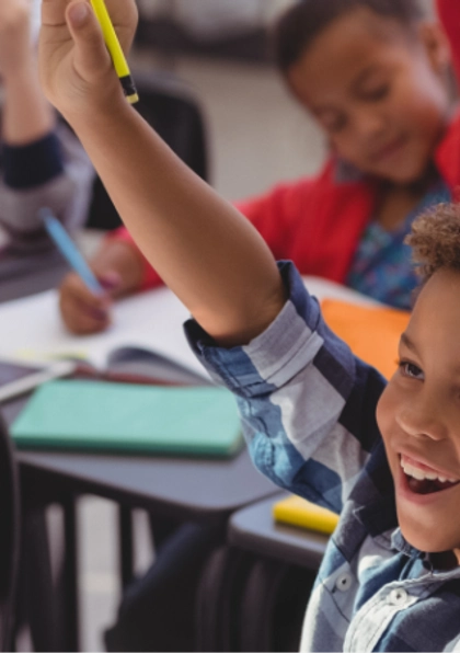 A group of young children in a classroom eagerly raise their hands, smiling and looking engaged. Notebooks and school supplies are visible on their desks.
