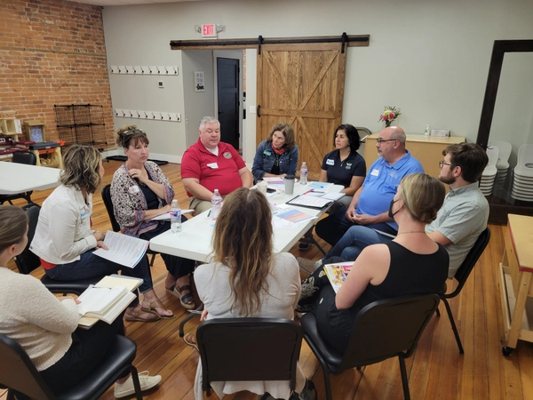 Eight adults sit in a circle around a table in a meeting room, engaged in discussion. Papers, notebooks, and water bottles are on the table. The space has wooden floors, brick walls, and a sliding barn door.