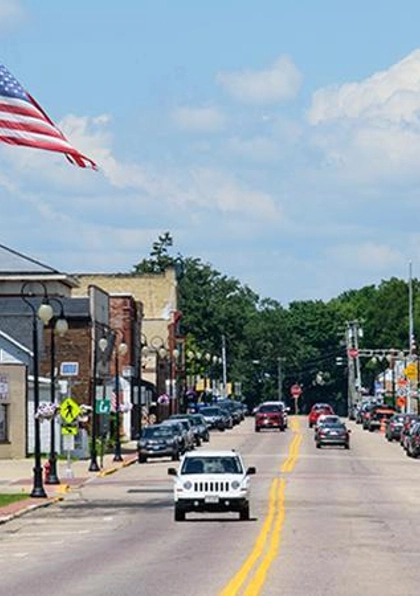 A small town main street lined with cars, brick buildings, shops, and an American flag flying on a pole. The sky is blue with some clouds, and people are visible on the sidewalks.