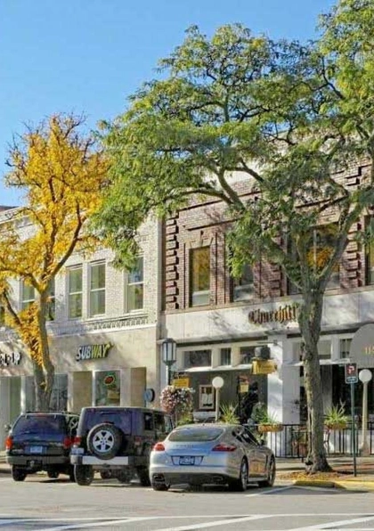 A sunny street lined with parked cars, trees with yellow and green leaves, and various storefronts including restaurants and shops in a small downtown area.