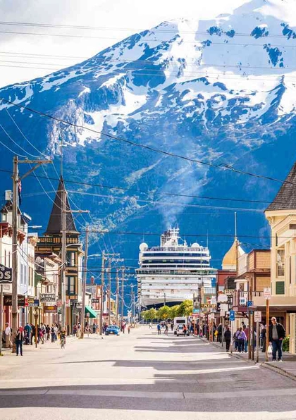 A bustling small-town street lined with colorful shops and people walking, with a large cruise ship docked at the end of the street and snow-capped mountains towering in the background.