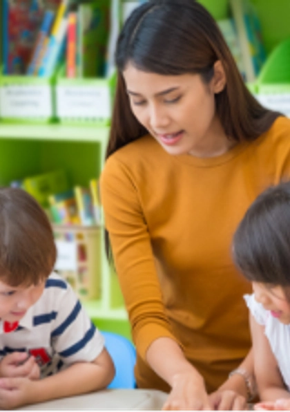 A teacher sits at a table with six young children in a colorful classroom, engaging them in an activity or lesson. Shelves filled with books and storage bins are visible in the background.