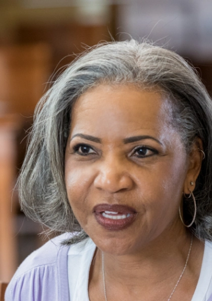 A middle-aged woman with gray hair, wearing hoop earrings and a light purple top, sits indoors and speaks, with a blurred background of wooden furniture.