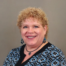 A smiling woman with short curly blonde hair wearing blue patterned earrings, a matching necklace, and a blue and black patterned top, posed in front of a plain beige background.