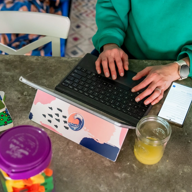 A caregiver types on a tablet keyboard at a table with children's books and a drink nearby.