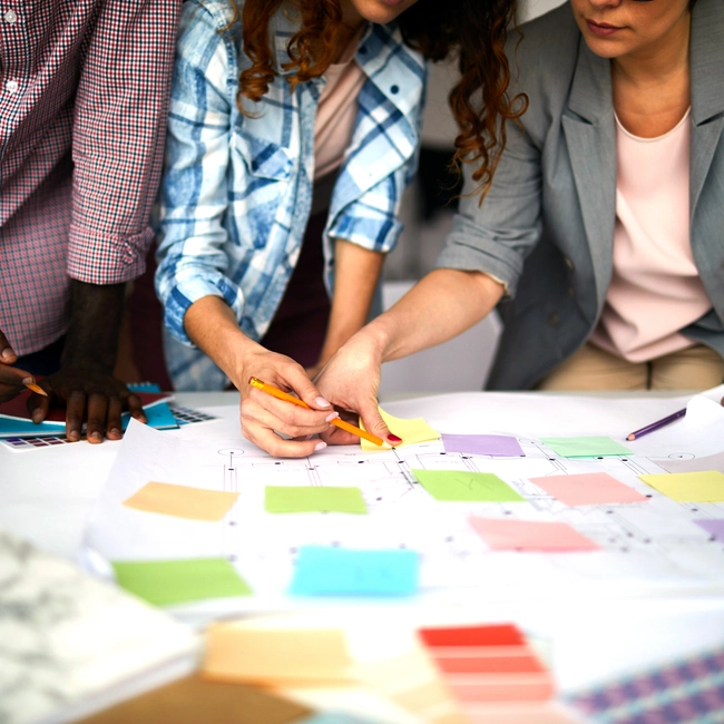 Three colleagues lean over a table covered in colorful sticky notes and planning documents.